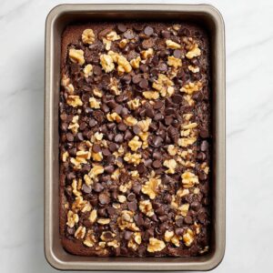 Best Chocolate Date Cake Recipe - Even Better Next Day 18 Top-down view of a baked chocolate date cake in a rectangular pan, topped with chocolate chips and chopped walnuts, light marble background, natural lighting, clean food photography.