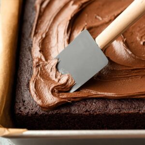 Easy Chocolate Mayonnaise Cake | 45-Minute Recipe 18 A close-up view of a spatula spreading smooth chocolate frosting over a freshly baked cake in a baking pan lined with parchment paper.