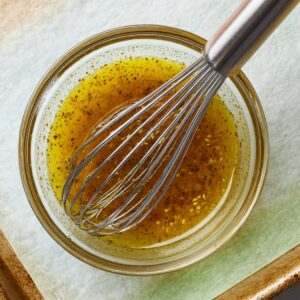 Christmas Salmon Recipe | Ready in 40 Minutes 15 “Top-down shot of a small bowl with vinaigrette-style sauce being whisked, visible olive oil and mustard texture, metal whisk, parchment-lined baking tray partially visible, soft natural lighting, clean recipe process photography.”