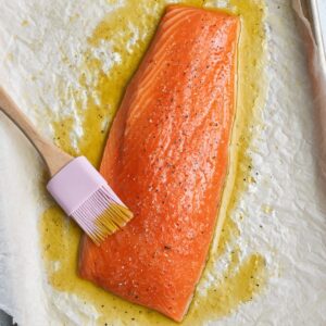 Christmas Salmon Recipe | Ready in 40 Minutes 16 “Overhead shot of raw salmon fillet placed on parchment-lined baking tray, brushed with olive oil and seasoning, pastel spatula on the side, minimal modern kitchen styling, neutral textured surface, clean and bright food photography, preparation stage.”
