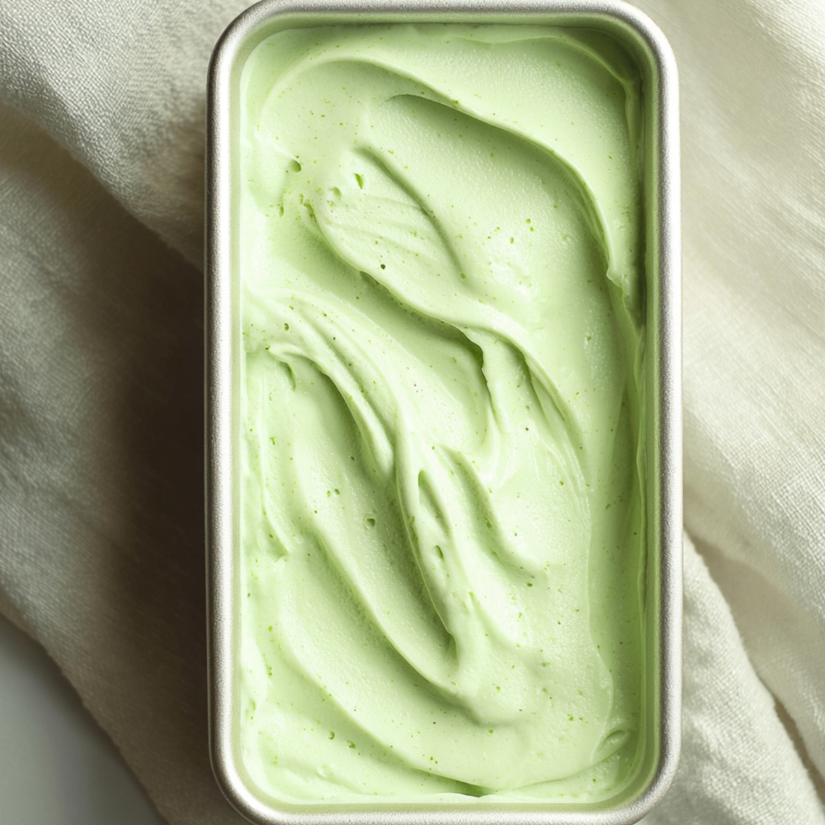 Grasshopper Drink Recipe | 4-Ingredient Frozen Cocktail 12 A rectangular container filled with a smooth, pale green frozen grasshopper drink, viewed from above on a light fabric background.