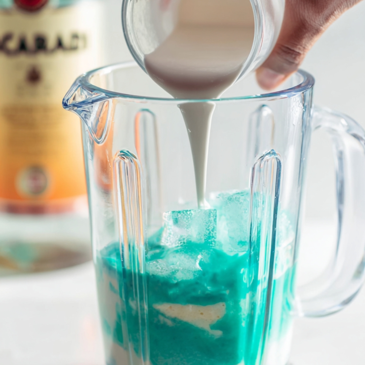 Jack Frost Cocktail Recipe | 5-Minute Frozen Drink 11 A hand pouring cream into a blender containing ice and a blue cocktail mixture, with Bacardi rum in the background