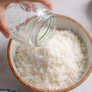 Jack Frost Cocktail Recipe | 5-Minute Frozen Drink 15 A hand pouring shredded coconut from a clear jar into a bowl filled with more shredded coconut.