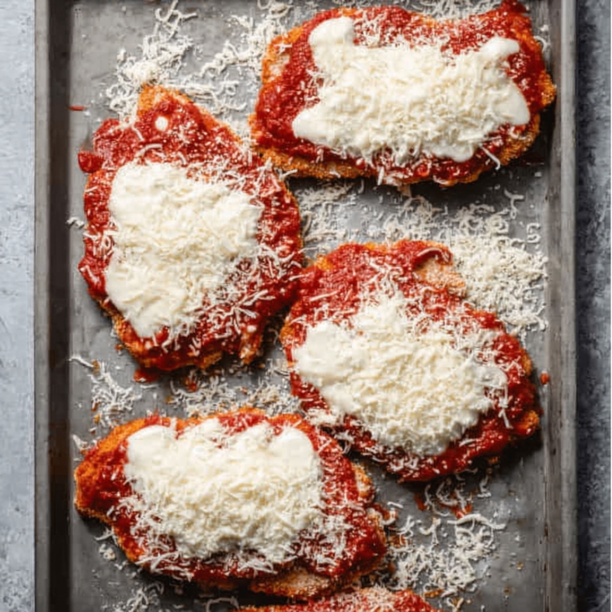 Easy Keto Chicken Parmesan Recipe 12 Raw chicken cutlets topped with tomato sauce and shredded mozzarella and parmesan cheese arranged on a baking tray before cooking.
