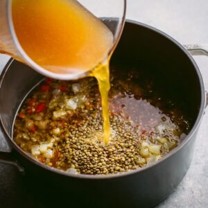 Best Lebanese Lentil Soup Recipe (Healthy & So Comforting!) 14 Pouring vegetable broth into a pot filled with dry lentils and chopped vegetables, ready for cooking.