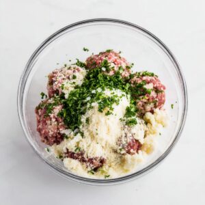 Easy Mozzarella Stuffed Meatballs | 8 Simple Steps (55 characters) 16 Raw meatball mixture in a glass bowl with ground beef, breadcrumbs, chopped onion, parsley and cheese, overhead food photography, bright natural light, clean white background, high detail