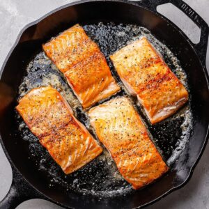 Salmon fillets searing in a cast iron skillet, lightly seasoned and browning on the surface.