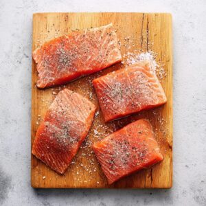 Raw salmon fillets seasoned with salt and black pepper on a wooden cutting board.