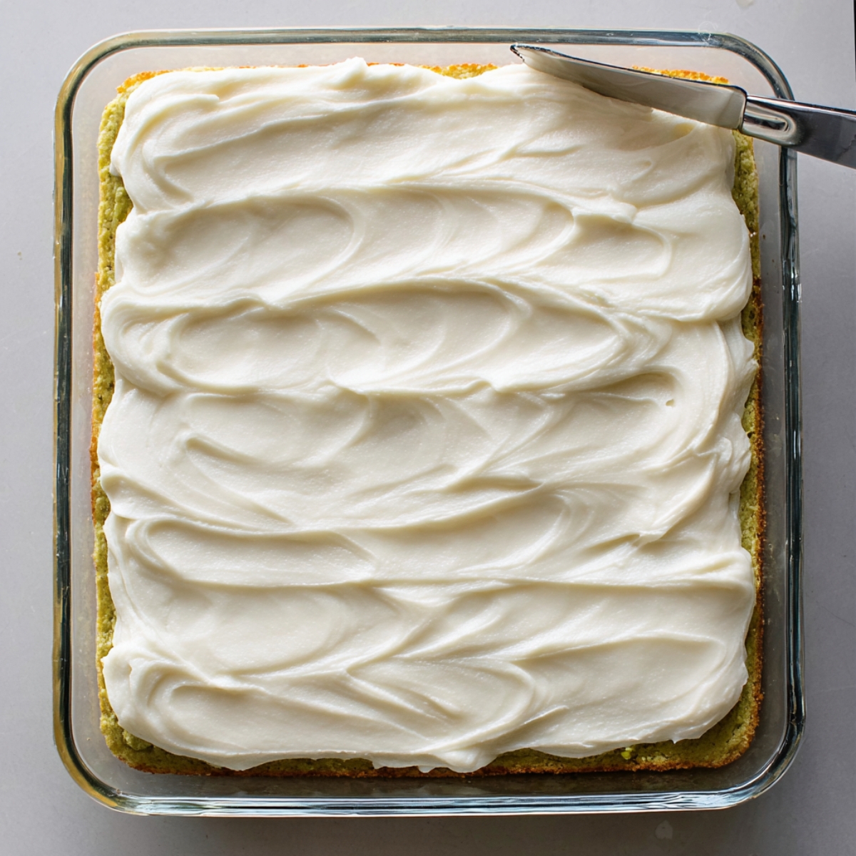 Easy Pistachio Cookie Bars | 40-Minute Dessert Recipe 12 Overhead shot of pistachio cookie bars in a clear glass baking dish, smooth white frosting spread evenly with visible spatula swirls, soft green base underneath, clean neutral background, bright natural lighting, modern baking photography style.
