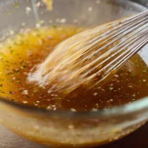 Best Red Wine Vinaigrette Recipe (Homemade in Minutes) 14 Close-up of red wine vinaigrette being whisked in a glass bowl, showing the dressing emulsifying with visible herbs and chopped shallots.