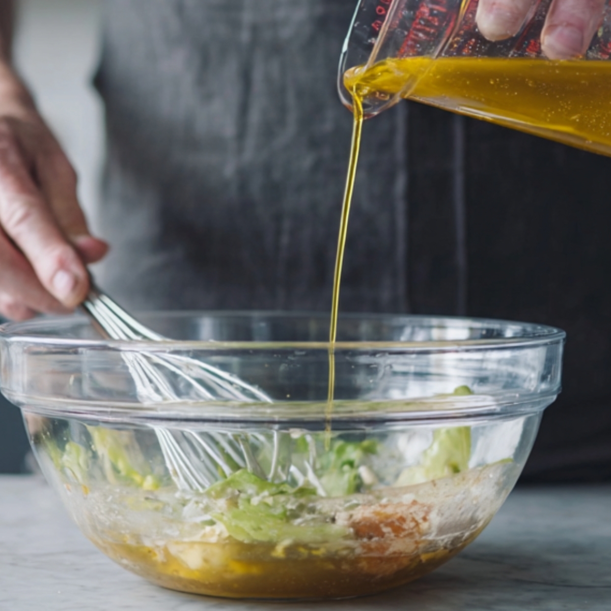 Best Red Wine Vinaigrette Recipe (Homemade in Minutes) 10 Olive oil being poured from a measuring cup into a glass bowl while whisking salad dressing during red wine vinaigrette preparation.