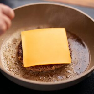 Smash Burger Tacos Recipe | Crispy & Easy in Minutes 17 A slice of cheese being placed on top of a sizzling beef patty in a pan, melting to create a gooey topping for a smash burger.