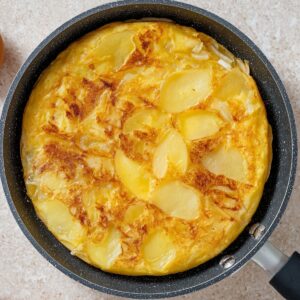 Top view of a Spanish tortilla cooking in a nonstick pan, sliced potatoes and onions set in eggs, golden yellow tones, minimal kitchen background, natural light, realistic food photography