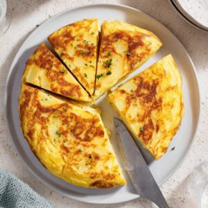 Overhead shot of a classic Spanish tortilla cut into wedges on a white plate, golden potato omelette, natural sunlight, Mediterranean food photography, clean neutral background