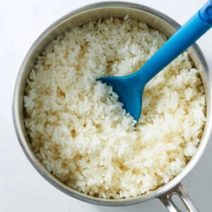 Stuffed Pepper Soup Recipe | Easy & Comforting in 52 Min 17 A top-down view of freshly cooked white rice in a stainless steel saucepan, fluffy and steaming, stirred with a blue silicone spoon, clean white background, natural soft lighting, food photography, high detail, realistic, minimal kitchen style