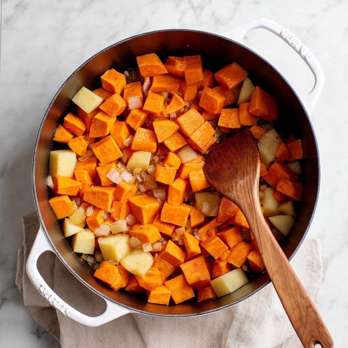 Easy Sweet Potato Soup Recipe | Vegan & Gluten-Free 10 “Top view of a pot filled with chopped sweet potatoes and potatoes being sautéed with onions, wooden spoon mixing, white enameled pot, clean marble surface, bright natural light, realistic food photography, high resolution”