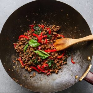 Thai Basil Beef Recipe - Quick, Easy & Ready in 25 Min 16 Wok filled with ground beef, red bell peppers, and fresh basil leaves being stirred with a wooden spoon