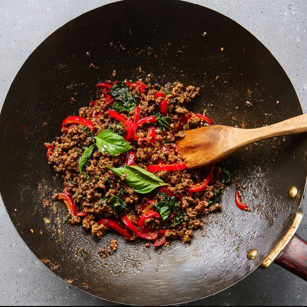 Thai Basil Beef Recipe - Quick, Easy & Ready in 25 Min 12 Wok filled with ground beef, red bell peppers, and fresh basil leaves being stirred with a wooden spoon