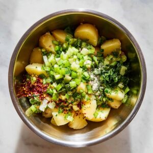 Turkish Potato Salad (No Mayo!) | 6 Easy Steps 15 Top-down view of a metal mixing bowl containing boiled potatoes topped with chopped green onions, parsley, red pepper flakes, salt, and olive oil before mixing.
