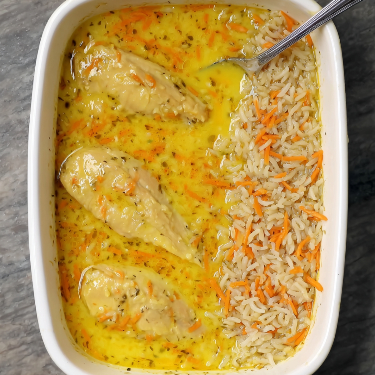 Healthy Turmeric Chicken and Rice Casserole 11 A casserole dish where rice and shredded carrots have been added to a golden, aromatic sauce. The chicken breasts are nestled within the dish, likely marinating in the flavorful liquid, and the rice is absorbing the vibrant color of the turmeric.
