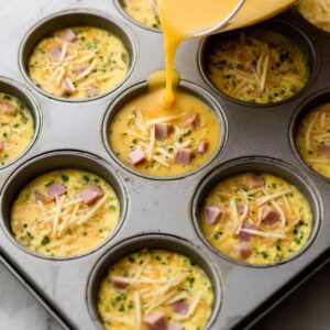 Healthy Breakfast Egg Muffins Recipe 17 A muffin tin filled with egg mixture, diced ham, and shredded cheese, ready to be baked. The egg mixture is poured over the ingredients in each muffin cup, awaiting cooking.