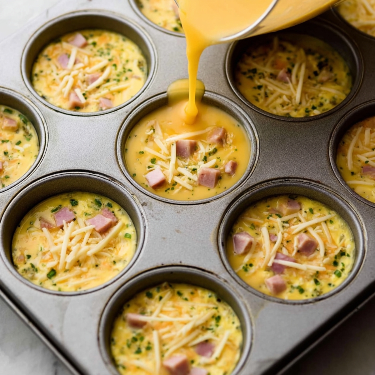 Healthy Breakfast Egg Muffins Recipe 12 A muffin tin filled with egg mixture, diced ham, and shredded cheese, ready to be baked. The egg mixture is poured over the ingredients in each muffin cup, awaiting cooking.