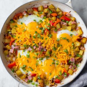 30-Minute Breakfast Skillet | Crispy Potatoes & Eggs 17 Breakfast skillet with baked eggs, melted cheddar cheese, roasted potatoes, ham, bell peppers, and green onions in a white pan.