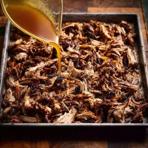 Carnitas Crockpot Recipe | 10 Ingredients, 8 Hours 17 Top-down view of shredded pork carnitas spread on a metal baking tray, crispy dark bits mixed with tender meat, rich pork juices being poured over the meat, rustic kitchen setting, warm lighting, realistic food photography, high resolution