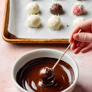 The Best Chocolate Covered Raspberries Recipe 14 White chocolate coated raspberries being dipped into melted dark chocolate with a skewer, overhead food photography on a pastel pink surface.