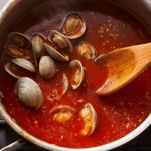 Clams gently added to the simmering tomato broth, their shells partially open as they cook, releasing their briny essence into the rich sauce. A wooden spoon stirs the mixture, blending the flavors.