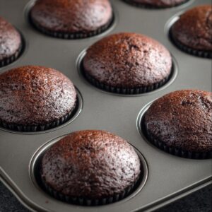 Easy Cream-Filled Chocolate Cupcakes | Homemade Recipe 16 Freshly baked dark chocolate cupcakes in a muffin tin, showing a shiny, domed top with slightly dark brown color, uniced and ready for frosting.