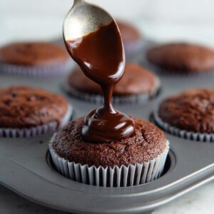 Easy Cream-Filled Chocolate Cupcakes | Homemade Recipe 18 A spoonful of thick dark chocolate ganache being drizzled onto a chocolate cupcake, with other cupcakes in the background.