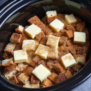 The Best Crockpot French Toast Recipe 17 A close-up shot of a slow cooker filled with cubed bread, topped with butter cubes and a generous sprinkle of cinnamon sugar. The bread is ready to be soaked in the sweet mixture and cooked into a delicious French toast casserole.