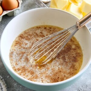 The Best Crockpot French Toast Recipe 16 A bowl with a cinnamon-sugar and egg mixture being whisked together with a metal whisk. The mixture is bubbly and ready to be poured over cubed bread for slow-cooker French toast casserole, with ingredients like eggs and butter visible in the background