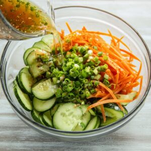 Healthy Cucumber and Carrot Salad Recipe 18 A glass bowl containing sliced cucumbers, julienned carrots, and chopped green onions, with a splash of dressing added for an Asian-style salad preparation.