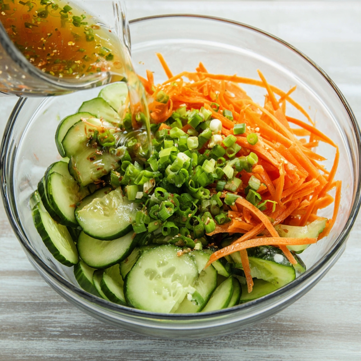 Healthy Cucumber and Carrot Salad Recipe 13 A glass bowl containing sliced cucumbers, julienned carrots, and chopped green onions, with a splash of dressing added for an Asian-style salad preparation.