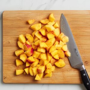 Brown Sugar Peach Cake Recipe | Easy & Moist 16 Freshly chopped peaches on a wooden cutting board with a chef’s knife, peach pieces lightly coated with spices on a marble surface.