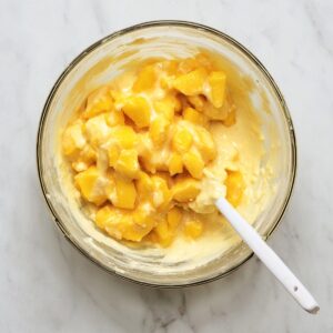 Brown Sugar Peach Cake Recipe | Easy & Moist 17 Overhead view of chopped peaches mixed into creamy cake batter in a glass bowl with a white spatula.