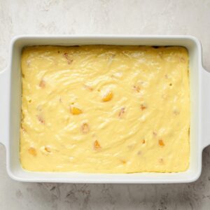Brown Sugar Peach Cake Recipe | Easy & Moist 18 Rectangular white baking dish filled with pale yellow cake batter dotted with peach pieces, viewed from above.