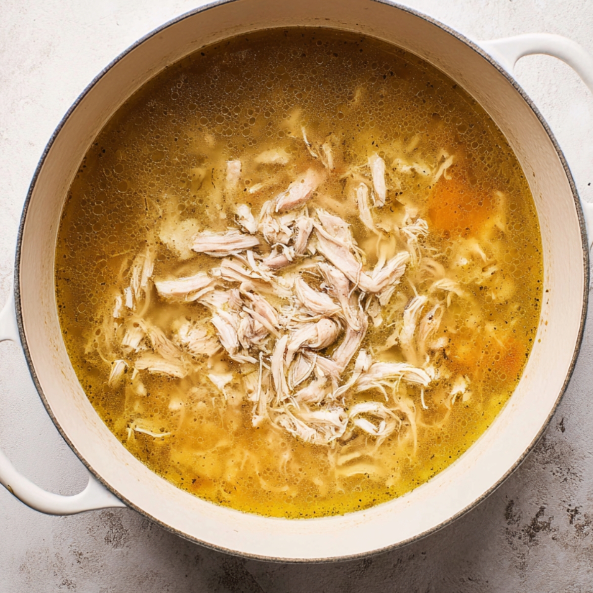 Ginger Chicken Soup Recipe | Easy & Comforting in 40 Min 11 Overhead view of a white pot filled with chicken broth and freshly shredded chicken, simmering during soup preparation.