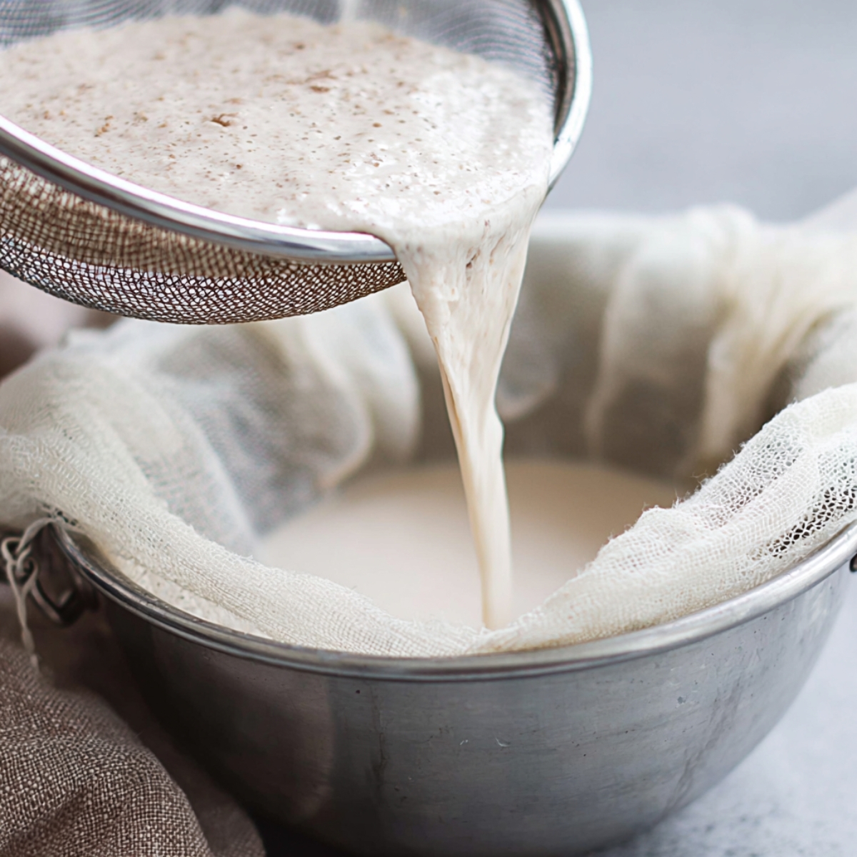 Authentic Horchata Recipe | Homemade Mexican Drink 12 Close-up food photography of homemade horchata being strained through white cheesecloth over a metal bowl, creamy rice milk flowing smoothly, natural soft daylight, minimal kitchen background, neutral tones, shallow depth of field, clean and rustic aesthetic, high detail, realistic texture, editorial food style.