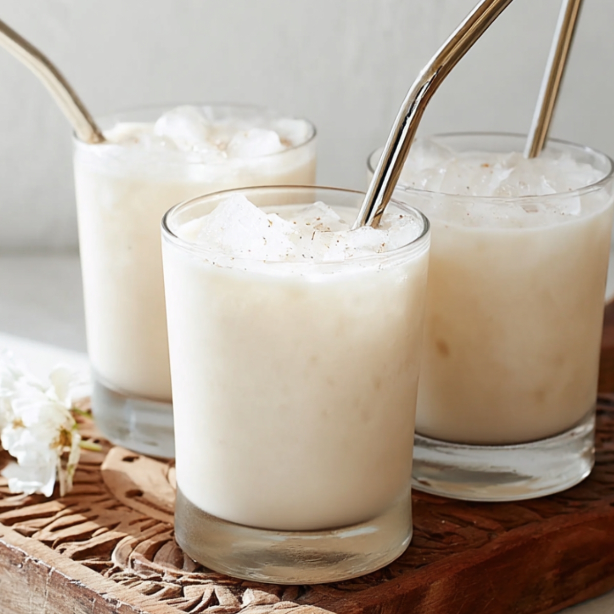 Authentic Horchata Recipe | Homemade Mexican Drink 14 Three small clear glasses filled with creamy Mexican horchata over ice, placed on a rustic carved wooden board, stainless steel reusable straws in the glasses, soft natural window light, white minimal background, elegant food photography, airy and bright mood, shallow depth of field, high detail, lifestyle drink photography, neutral tones, 50mm lens, f/2.8
