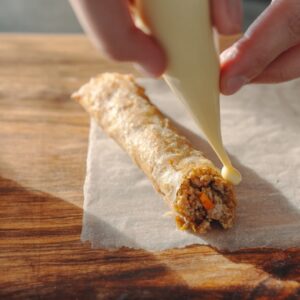 Crispy Lumpia Recipe | Easy Filipino Spring Rolls 15 Hands sealing a lumpia spring roll wrapper filled with meat and vegetables using a piping bag on a wooden surface