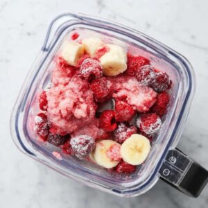 Raspberry smoothie bowl topped with granola, fresh berries, and banana slices, placed on a marble countertop.