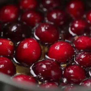 Cranberries simmering in a sugar syrup in a saucepan, appearing glossy and red.