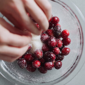 Overhead view of a hand sprinkling sugar over fresh cranberries in a glass bowl.