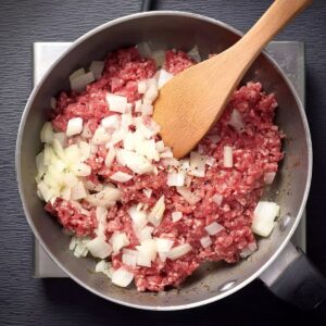 Taco Potatoes Recipe | Double Stuffed & Cheesy 16 Top-down food photography of raw ground beef and chopped onions cooking together in a saucepan, wooden spoon stirring, early cooking stage, natural kitchen light, realistic home cooking scene, instructional recipe photography, high realism