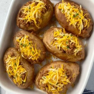 Taco Potatoes Recipe | Double Stuffed & Cheesy 17 Overhead food photography of baked potatoes stuffed with seasoned ground beef and topped with shredded cheddar cheese, arranged in a white baking dish, simple home-cooking style, soft kitchen lighting, realistic textures, casual comfort food photography, high detail