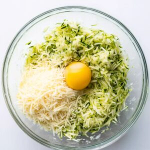 Zucchini Breadsticks Recipe | 5 Ingredients & Ready in 40 Min 15 Glass mixing bowl filled with grated zucchini, shredded cheese, and a whole egg before mixing for cheesy zucchini breadsticks