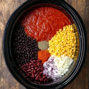 Easy Cheesy Taco Pasta Recipe 15 Ingredients for a slow-cooked cheesy taco pasta, including black beans, corn, diced onions, tomato sauce, and seasonings, all neatly placed in the slow cooker. The vibrant colors of the ingredients make the dish visually appealing.